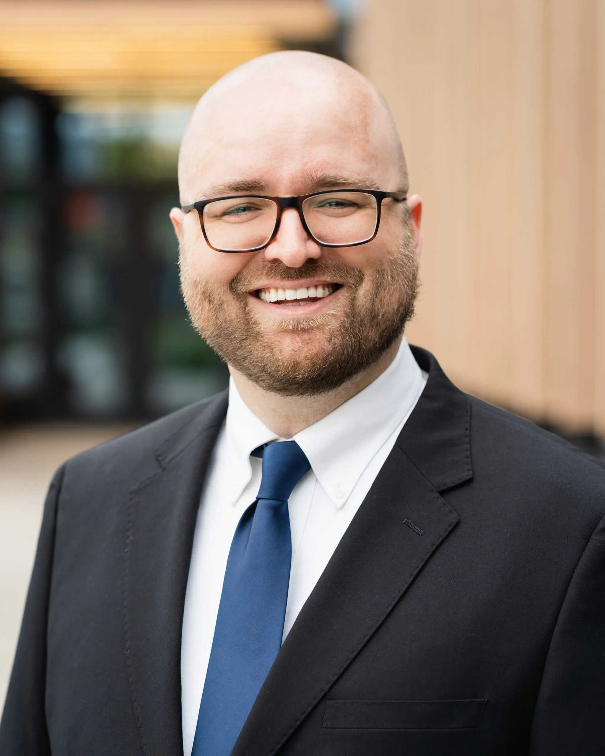 Ben Fisher, bald caucasian man with short dark beard. Wearing black rimmed glasses, dark suit with white shirt and blue tie. Smiling. 